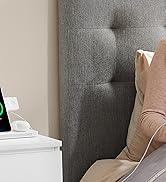 Dispositif d'affichage intelligent sur table de chevet blanche, affichant l'heure 9:41. Tête de lit capitonnée grise visible avec coussin beige. Vue partielle du câble de charge connecté à l'appareil.