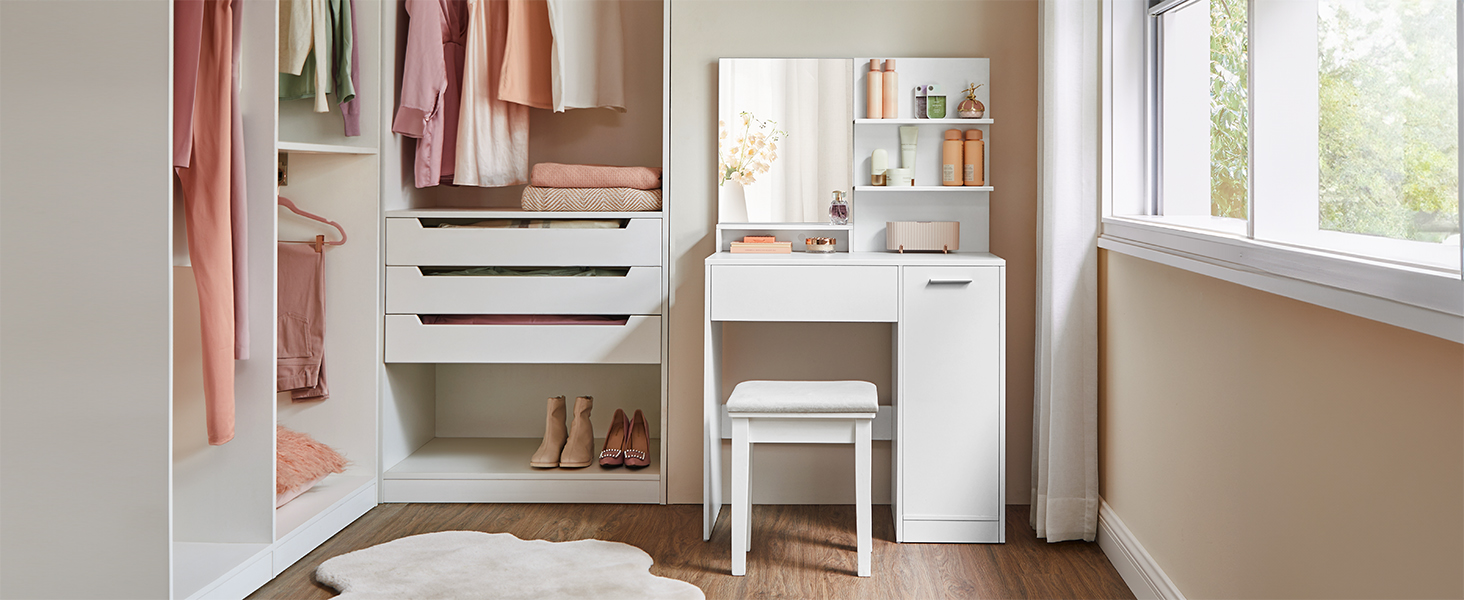 Espace de rangement organisé avec étagères blanches, tiroirs et tringles de suspension. Comprend une petite table de toilette blanche et un tabouret près d'une fenêtre.